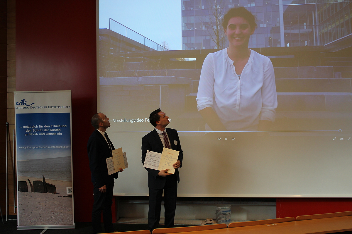 Zwei Gewinner des Förderpreises der Stiftung Deutscher Küstenschutz stehen vor einer Leinwand, eine Gewinnerin ist Live per Video zugeschaltet und auf der Leinwand zu sehen. 